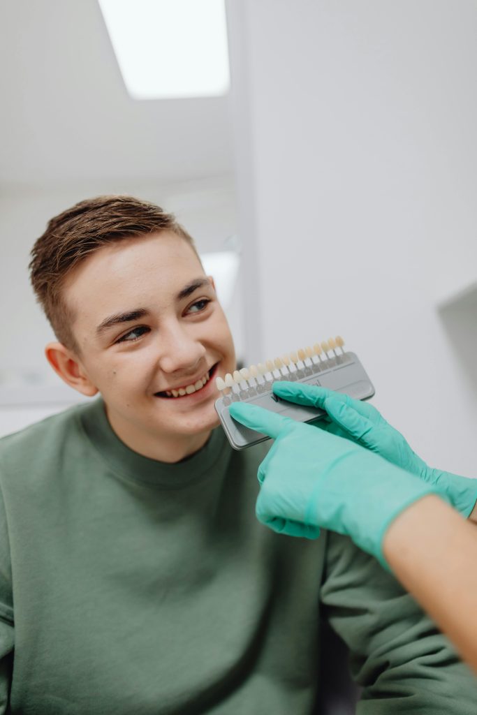 Smiling teenage boy at dental checkup with shade guide in hands of dentist wearing green gloves.